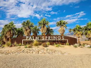 Palm-Springs-Schild zwischen Palmen vor Bergen und einem blau bewölkten Himmel.