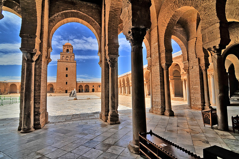 Historische Arkaden mit Blick auf Minarett und Innenhof unter blauem Himmel.
