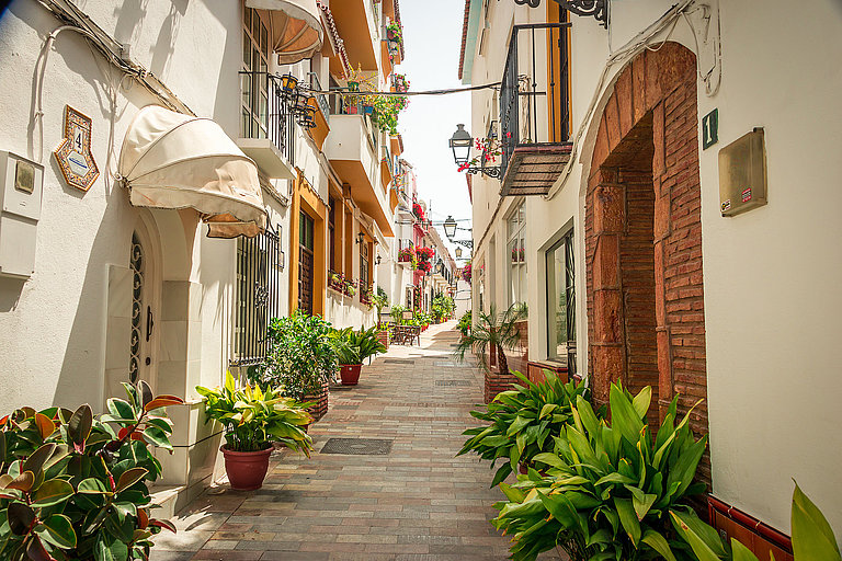 Sonnige Altstadtgasse in Marbella mit weißen Häusern, Pflanzen und Balkonen.