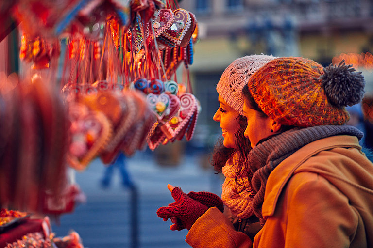 Das Bild zeigt zwei warm angezogene Frauen, die auf einem Weihnachtsmarkt die Lebkuchenherzen anlächeln.