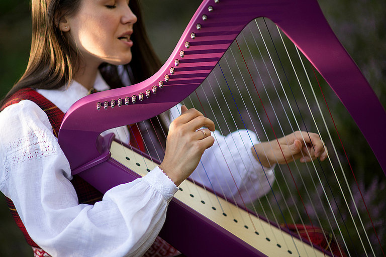 Junge Musikerin mit einer Harfe: Das Bild zeigt eine junge Frau, die eine Harfe spielt. Sie trägt ein traditionelles weißes Kleid mit einem roten Schal und ist in ihre Musik vertieft. Die Harfe hat einen glänzenden, violetten Rahmen, und die Harfensaiten werden von ihren Fingern sanft gezupft.