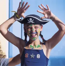 Mädchen mit bunter Gesichtsbemalung hebt lächelnd die Hände auf einem Kreuzfahrtschiff.