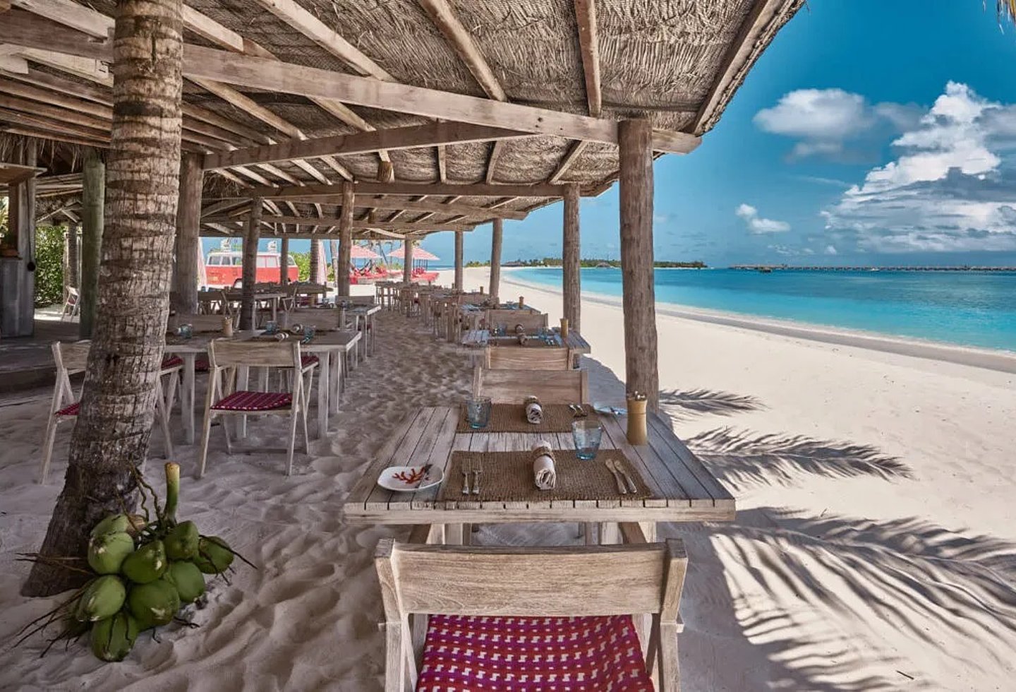 Überdachte Strandterrasse mit Tischen, Stühlen, Palmen und Blick aufs Meer.