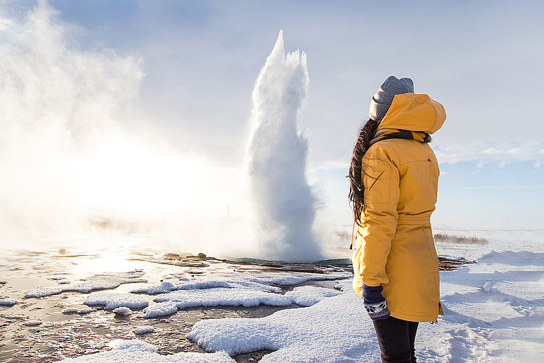 Person im gelben Mantel vor Geysirlandschaft im Winter unter dramatischen Himmel.