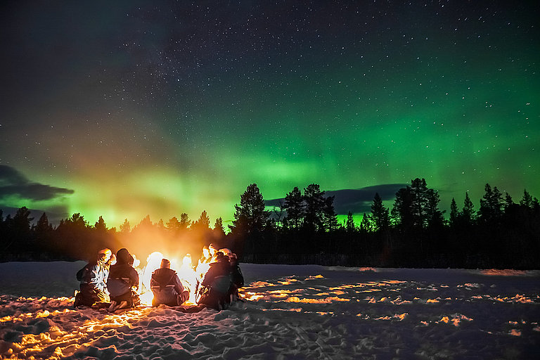 Lagerfeuer im Schnee unter Nordlichtern und Sternenhimmel vor dunklem Waldrand.