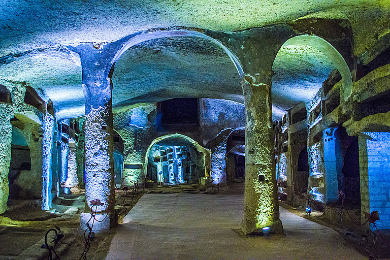 Gewölbehalle mit steinernen Säulen und blau beleuchteten Wänden im Untergrund.