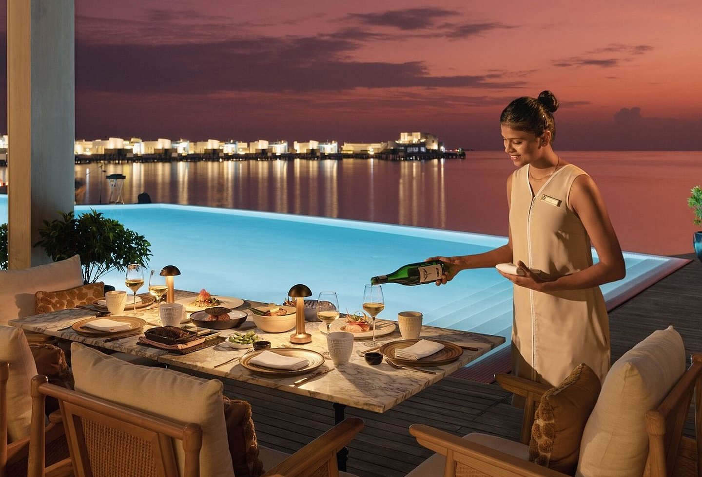 Mitarbeiter deckt einen Esstisch am Infinitypool bei tropischem Sonnenuntergang.