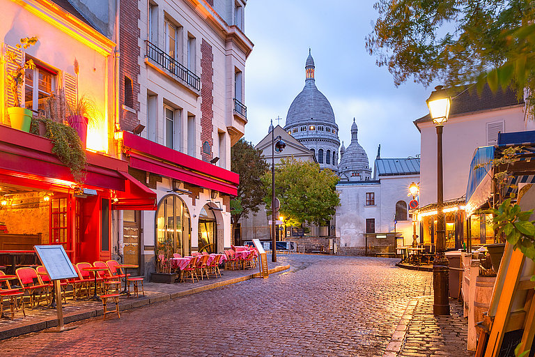 Beleuchtete Gasse in Montmartre mit Cafés und blick auf Sacré-Coeur am Abend.