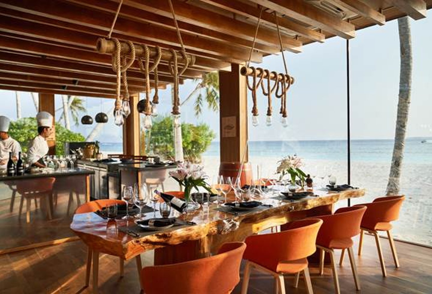 gedeckter Holztisch, orange Stühle, Glasfront mit Blick auf Strand und Meer, Buffet mit Köchen