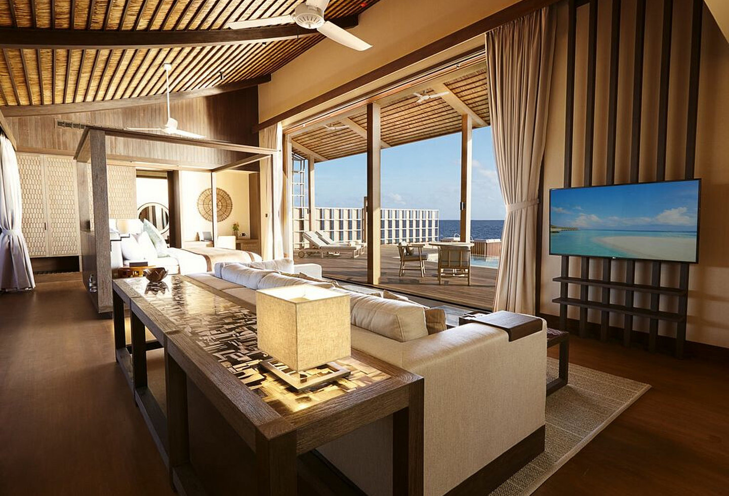 Blick über Couchbereich und TV zum Bett und auf die Terrasse mit Pool und Liegen