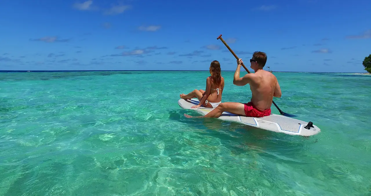 Zwei Menschen sitzen auf einem Surfboard und paddeln in der Lagune.