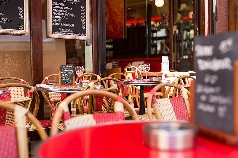 Charakteristisches Pariser Café: Bunte Stühle, Tische mit Menüs und einem Glas Wein – der perfekte Ort, um das Leben à la ‚Emily in Paris‘ zu genießen.