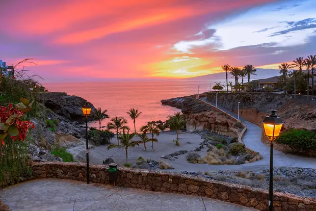 Strandbucht auf Teneriffa bei pinkem Himmel mit Palmen und Spazierweg entlang des Ufers.