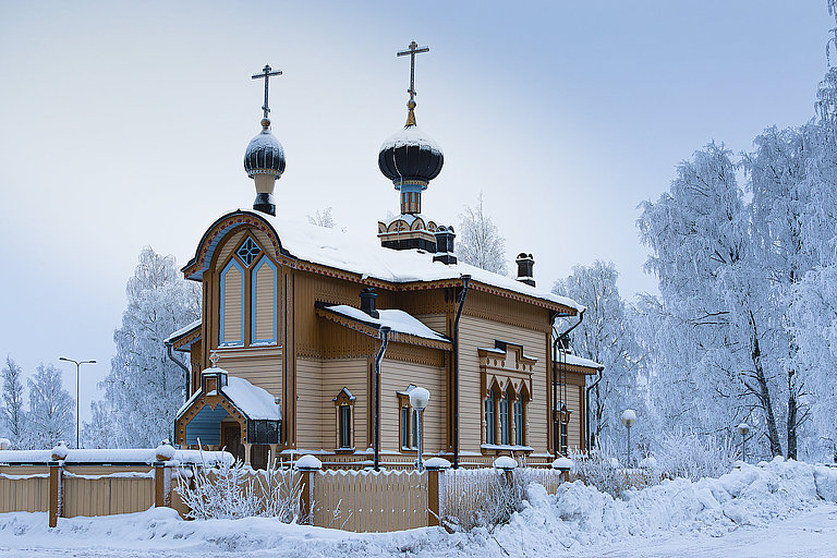 Verschneite Holzkirche mit Zwiebelturm in winterlicher Landschaft unter grauem Himmel.