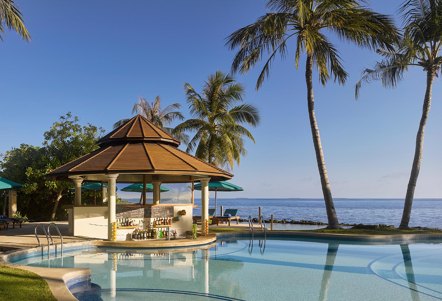 Pool mit Pavillon, Palmen und Blick auf das Meer unter strahlend blauem Himmel.