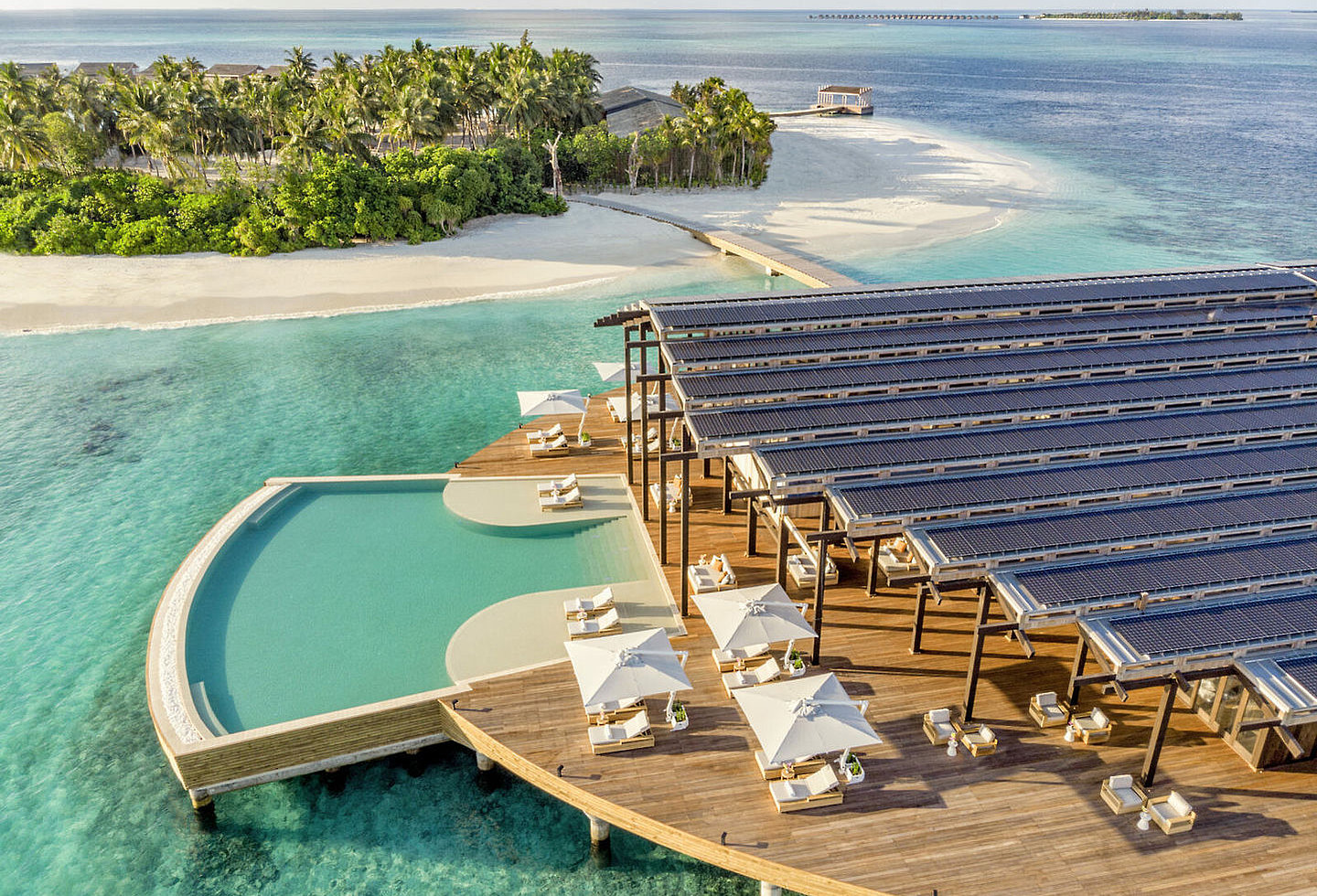 Blick auf Pool direkt über dem Meer, Holzterrasse mit Liegen und Schirmen, große Dachfläche mit Photovoltaik-Modulen