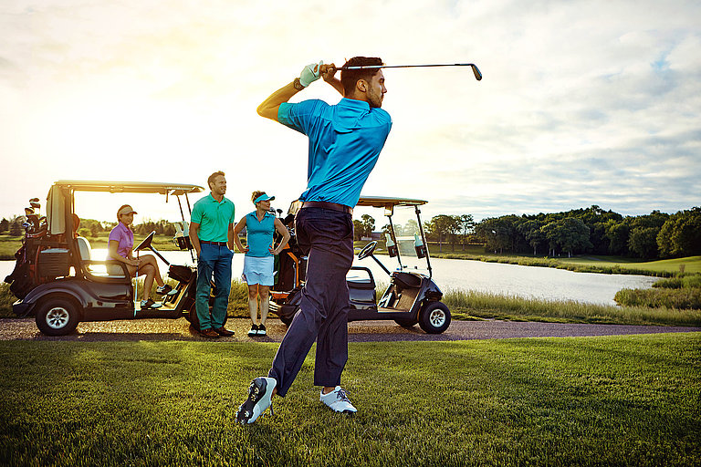 Golfer in bunter Kleidung beobachten einen Mann beim Abschlag mit blauem T-Shirt.
