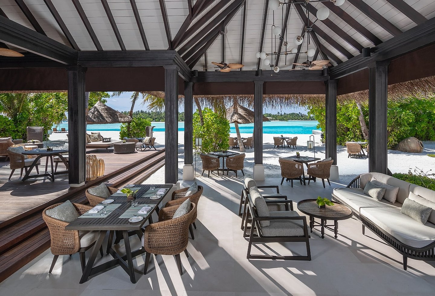 Offener Resortpavillon mit Sitzgruppen, Palmen und Blick auf Pool und Meer.