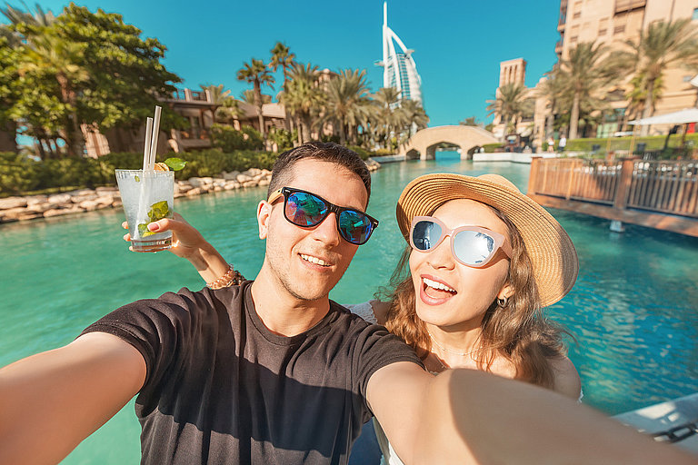 Zwei Touristen fotografieren sich mit Cocktails in der Hand, dahinter ein kristallklarer Fluss.