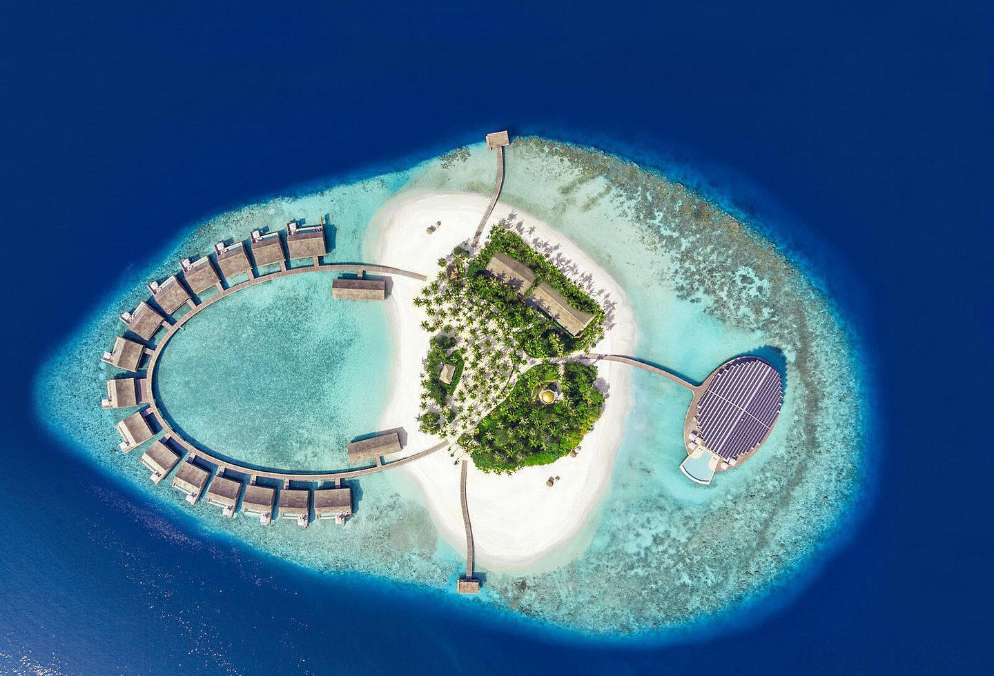 Vogelperspektive auf die kleine Insel mit viel Sandstrand, den Wasservillen und auf der anderen Seite futuristisches Gebäude und Holzterrasse mit Pool im Meer