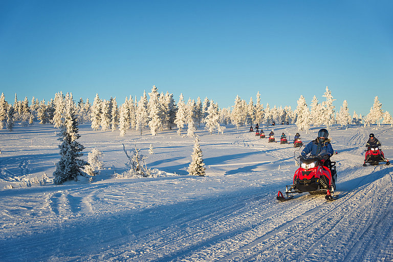 Zwei Schneemobile fahren durch eine verschneite Winterlandschaft unter blauem Himmel.
