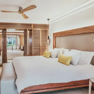 Helles Hotelzimmer mit Doppeltbett, Holzregal, Ventilator und Blick zum Meer.