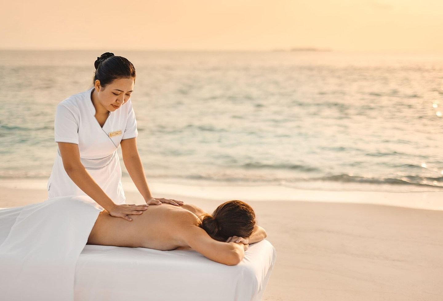Masseurin behandelt einen Gast am Strand vor ruhigem Meer im warmen Abendlicht.
