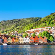 Bunte Häuser am Fjordufer in Bergen vor grünen Hügeln und blauem Himmel.