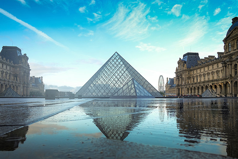 Gläserne Pyramide des Louvre spiegelt sich auf nassem Boden im Innenhof, umgeben von historischen Museumsgebäuden unter blauem Himmel.