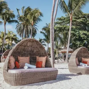 Strandbereich mit runden Strohdach-Liegen, Palmen und Kies vor blauem Meer.
