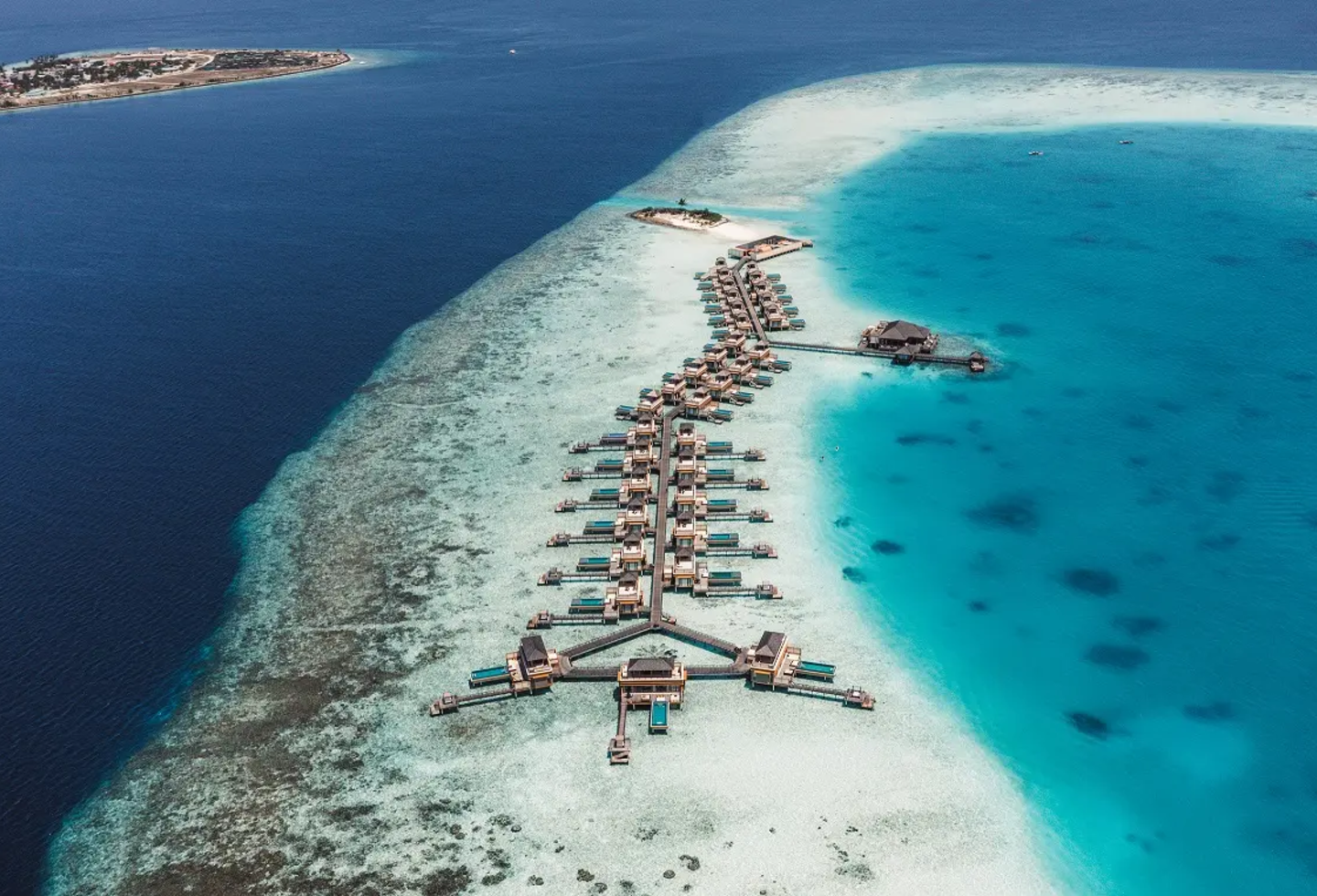 Luftaufnahme eines Korallenriffs mit Sandbank und Steg im türkisblauen Meer.