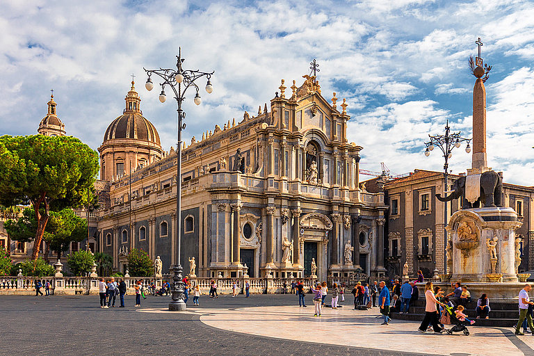 Hauptplatz in Catania, Sizilien mit Kathedrale, Passanten und blauem, leicht bewoelktem Himmel.