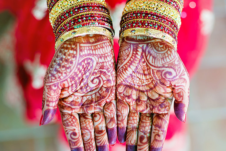 Mit Henna verzierte Hände mit buntem Armschmuck vor rotem Gewand im Hintergrund.