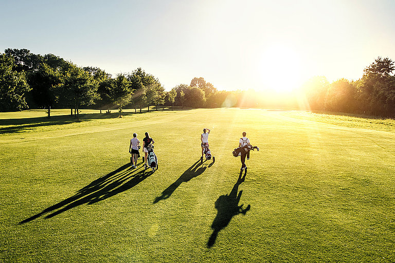 Vier Golfer, drei Frauen und ein Mann, gehen auf einem sonnigen Golfplatz. 