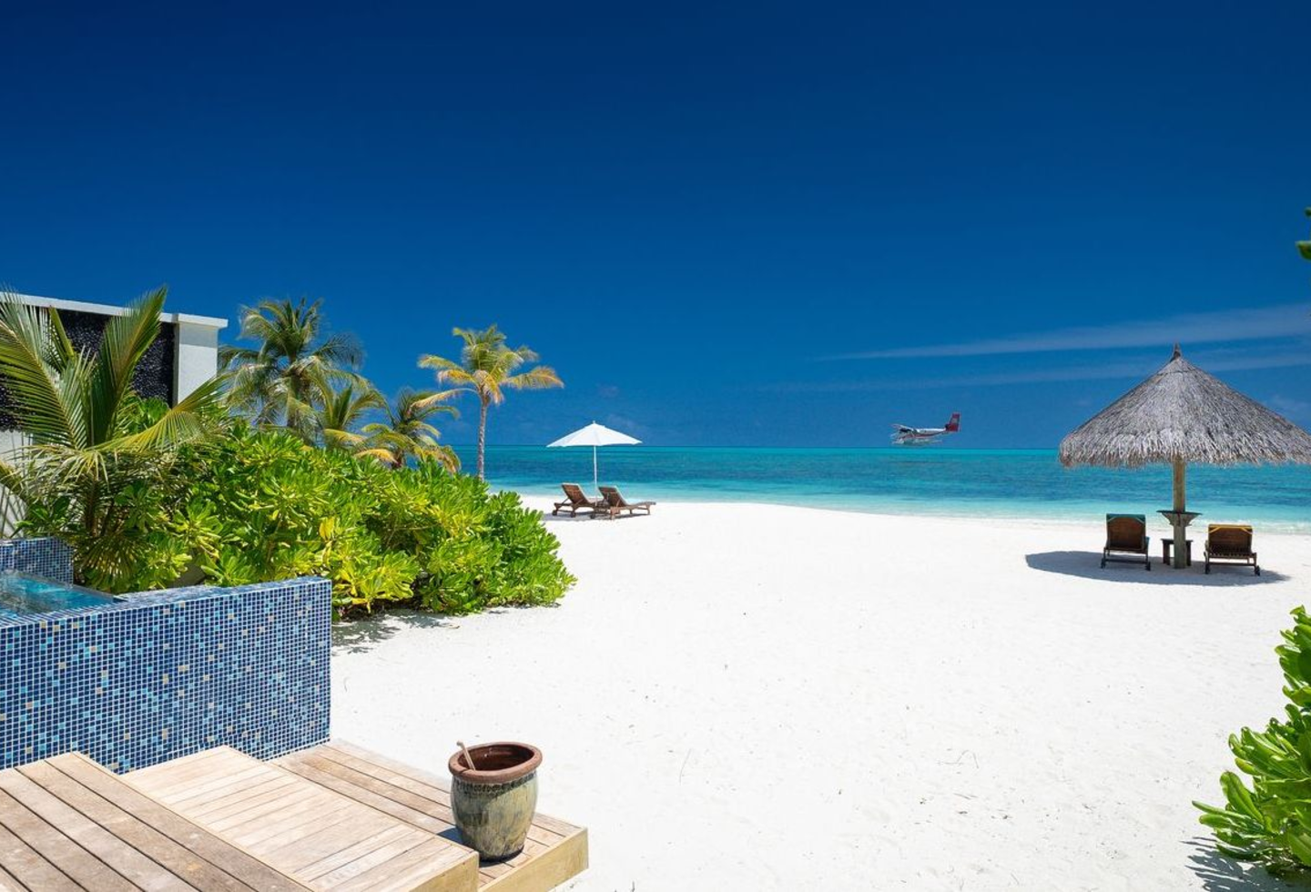 Weißer Sandstrand mit türkisblauem Meer, Palmen und einem Pavillon am Horizont.