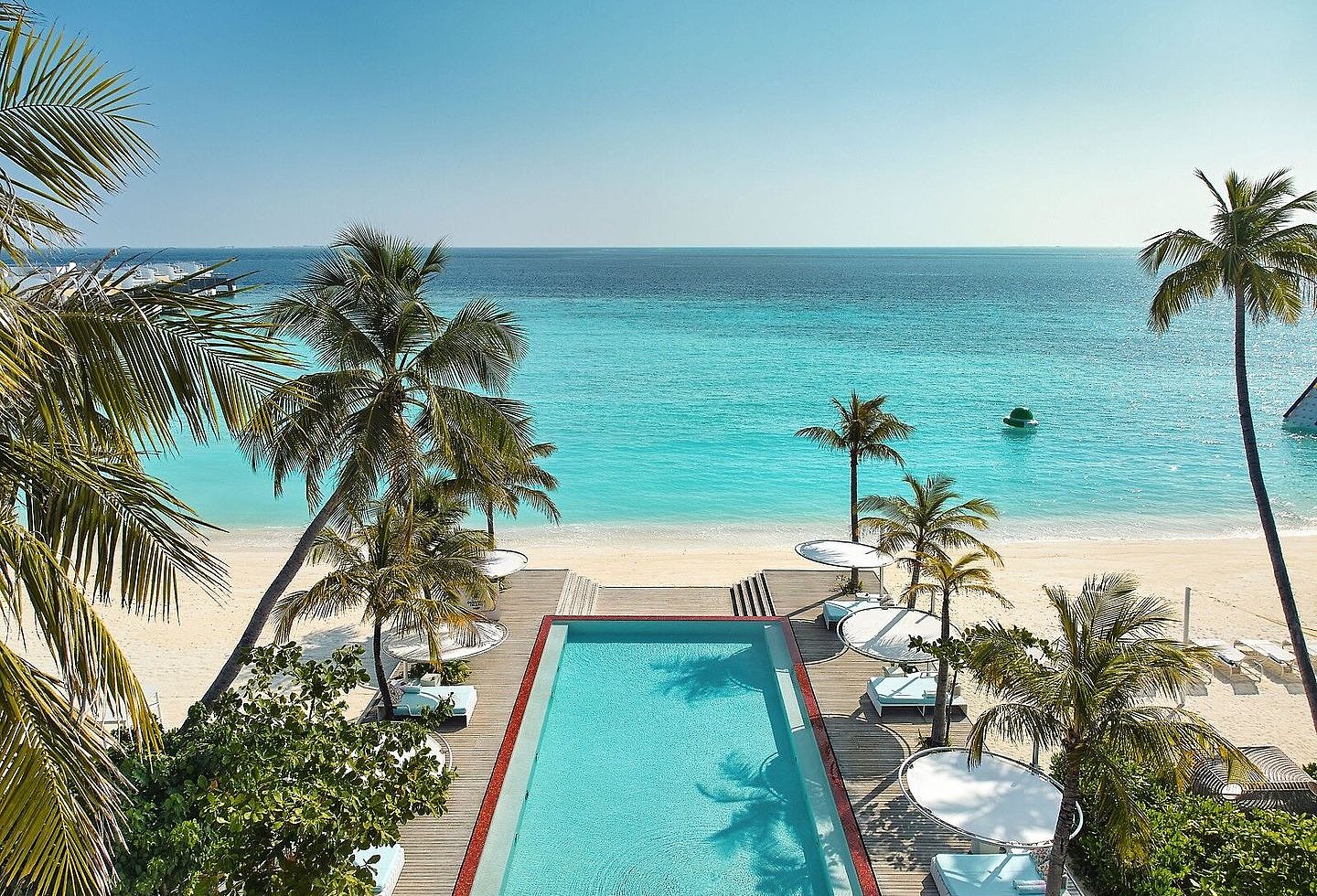 Blick vom Pool mit Holzterrassen, Liegen und Schirmen auf den weißen Sandstrand und das türkise Meer.