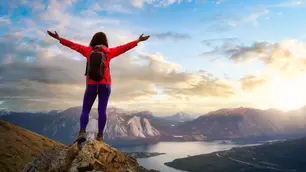 Person mit roter Jacke und Rucksack steht mit offenen Armen auf einem Felsen und blickt ins Tal.
