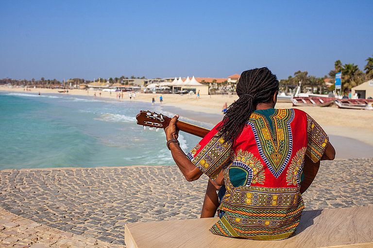 Das Bild zeigt einen Mann, der am Strand von Santa Maria auf der Insel Sal sitzt und Gitarre spielt. 