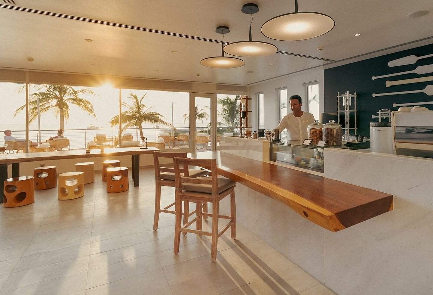 Helles Restaurant mit Holztischen, Bar und Blick auf Palmen und den Strand.