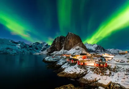 Grüne Nordlichter über verschneitem Lofoten-Dorf, Berg und dunkler Küste.