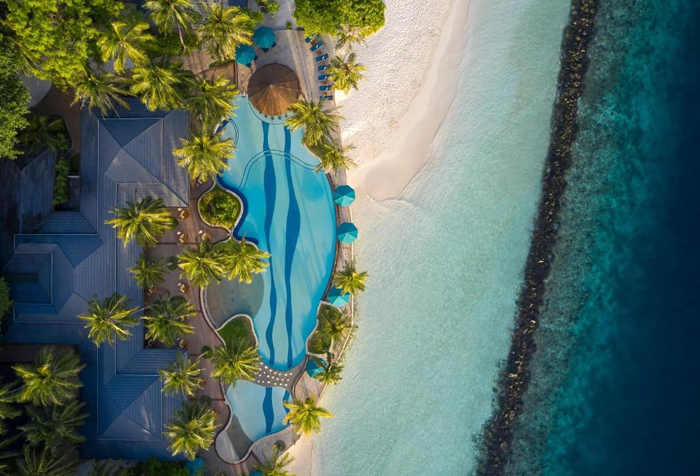 Luftaufnahme eines weißen Sandstrands mit Palmen, Liegen und türkisblauem Meer.