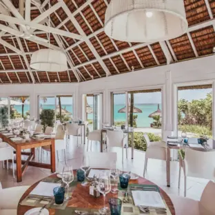 Helles Restaurant mit gedeckten Tischen, Korbstühlen und Blick auf das Meer.
