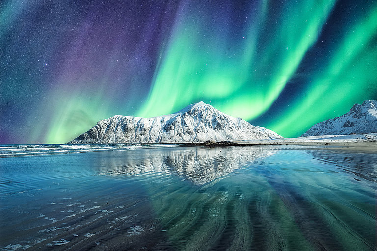 Grüne Nordlichter über verschneiter Küste, Bergen und spiegelndem Wasser.