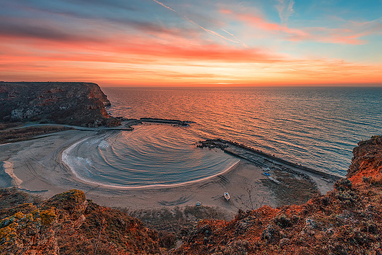 Bucht mit ruhigem Meer, Felsen und glitzerndem Wasser bei Sonnenuntergang.