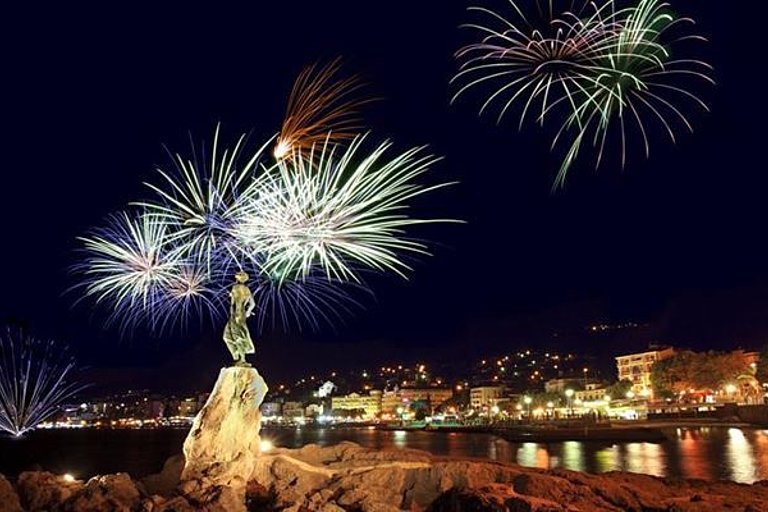 Feuerwerk über einer Stadt am Wasser mit Lichtern und nächtlicher Feststimmung.