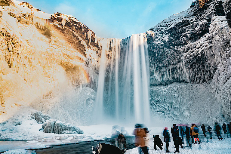 Hoher Wasserfall vor schneebedeckten Bergen, mit BesucherInnen am Aussichtspunkt.