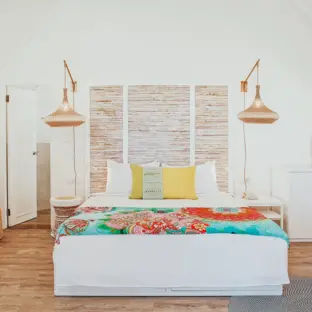 Helles Hotelzimmer mit buntem Bett, Wandteppich und orangefarbenen Hängelampen.